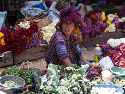 Marktvrouw met paarse hoofddoek tussen groenten en kleurrijke bloemen langs de Route Trans Midden Amerika.