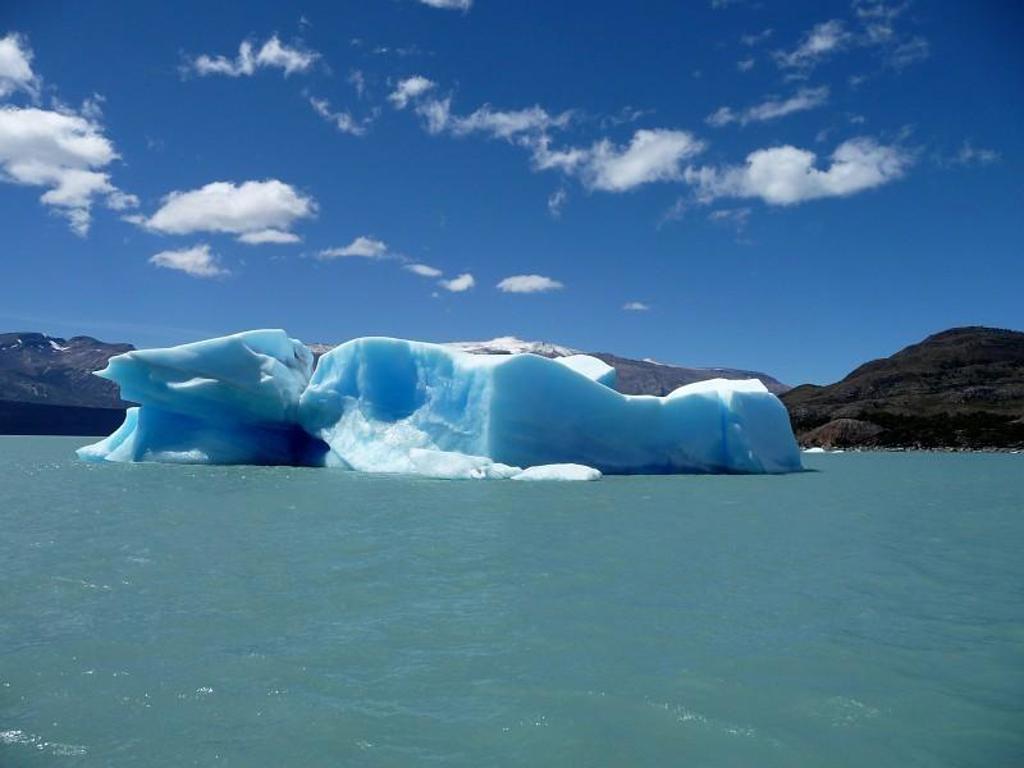 Blauwe ijsberg drijvend in een azuurblauw meer in Patagonië, met bergen op de achtergrond.