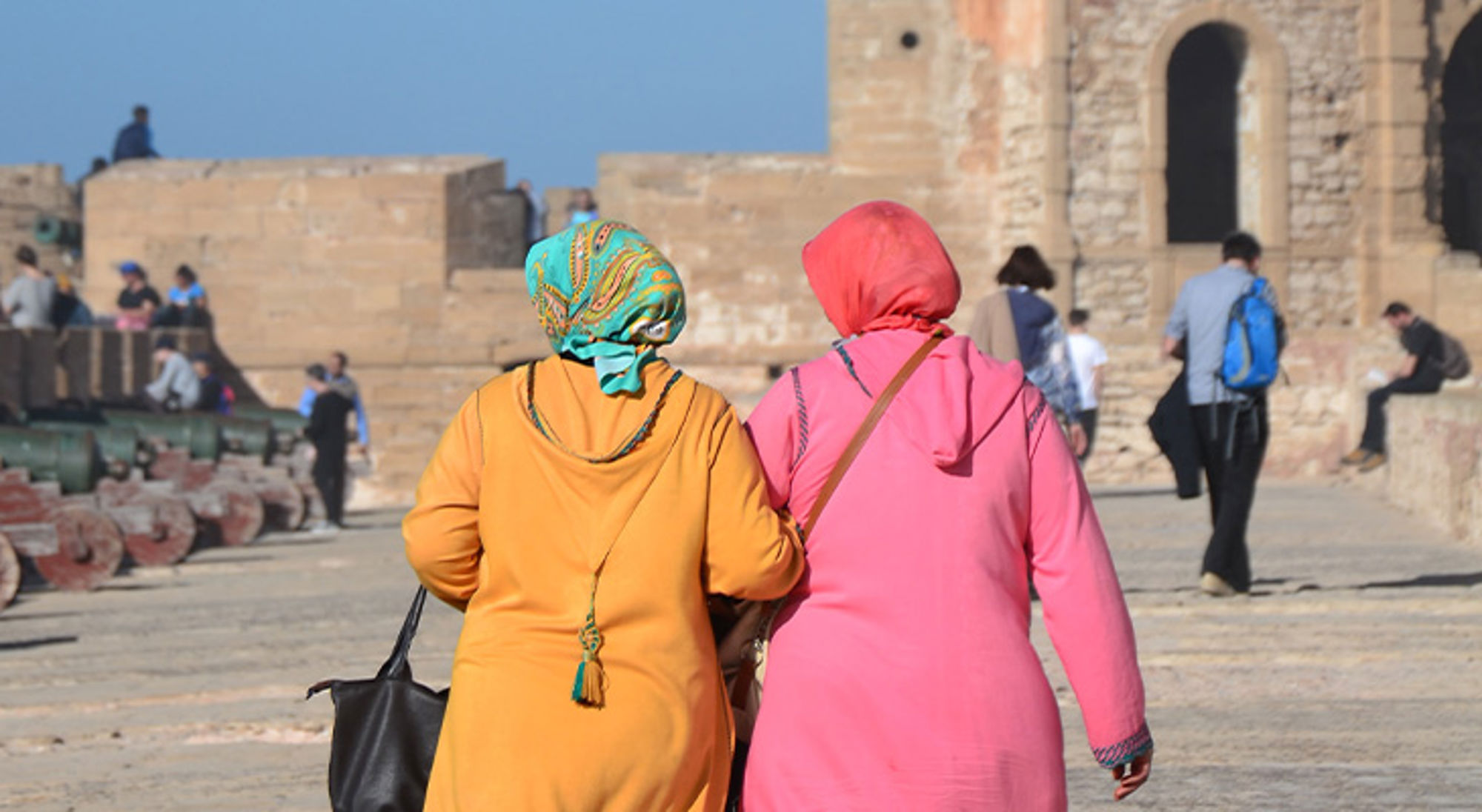 Islam in Marokko Twee vrouwen in kleurrijke kleding wandelen langs een historisch fort met andere bezoekers op de achtergrond.