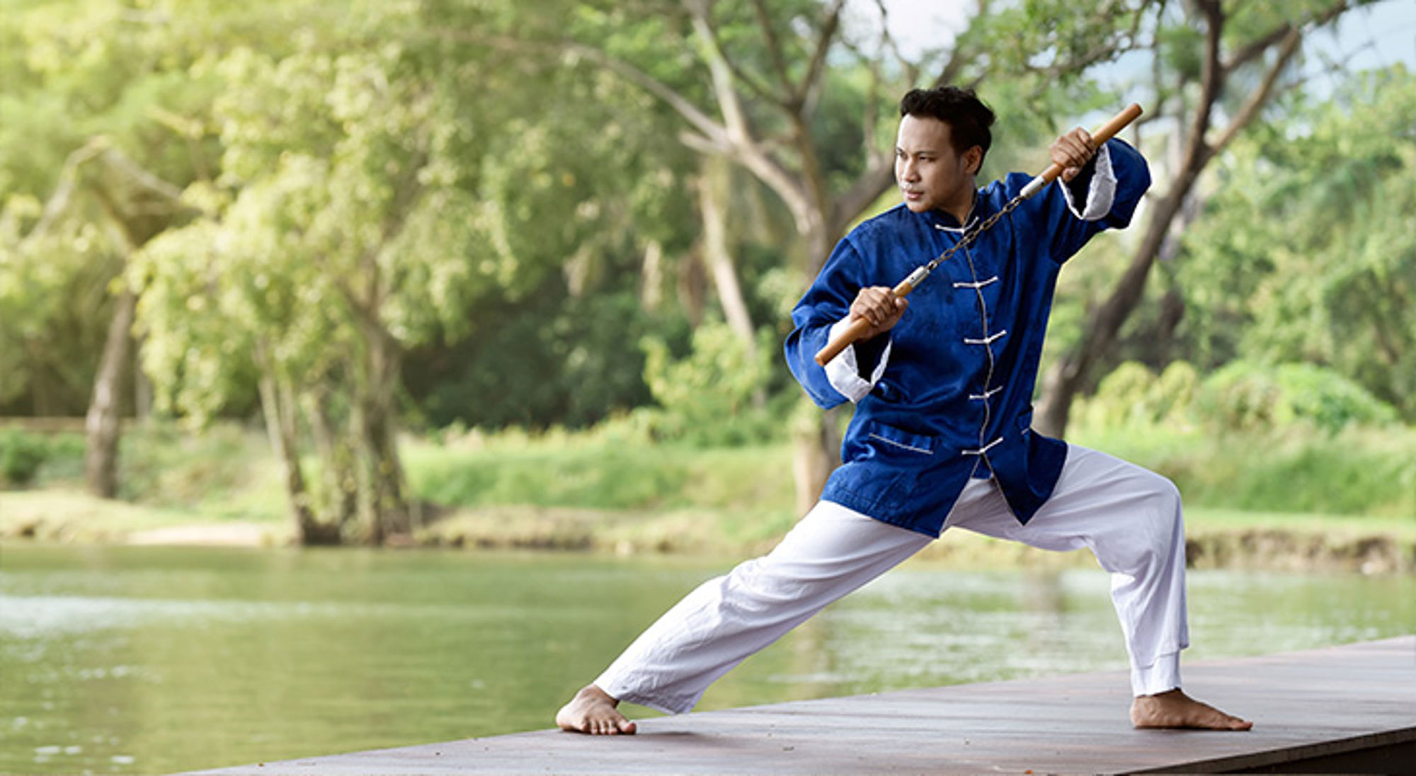 China Kung Fu Man beoefent kungfu met stok op een houten steiger aan het water, met groene bomen op de achtergrond.