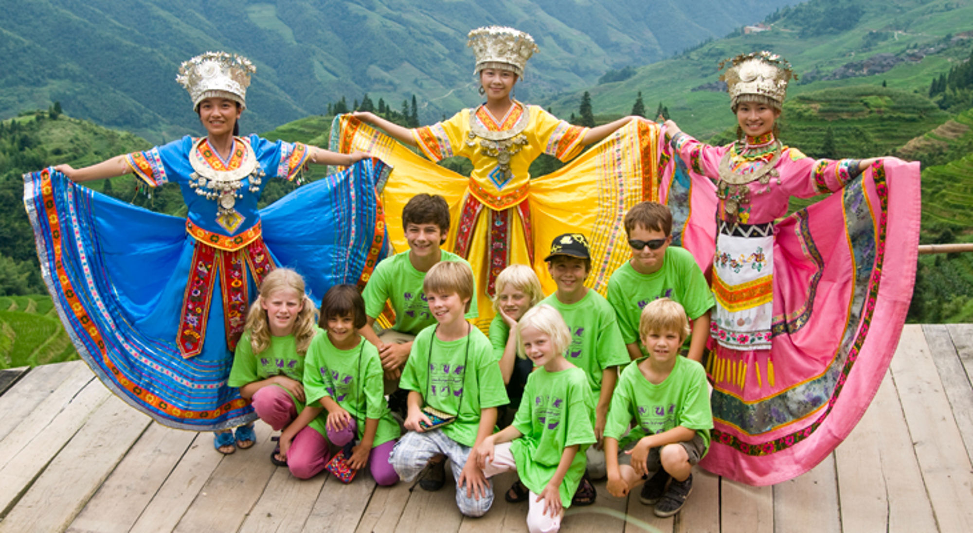 Kinderen poseren met drie vrouwen in kleurrijke traditionele klederdracht, met rijstterrassen op de achtergrond.