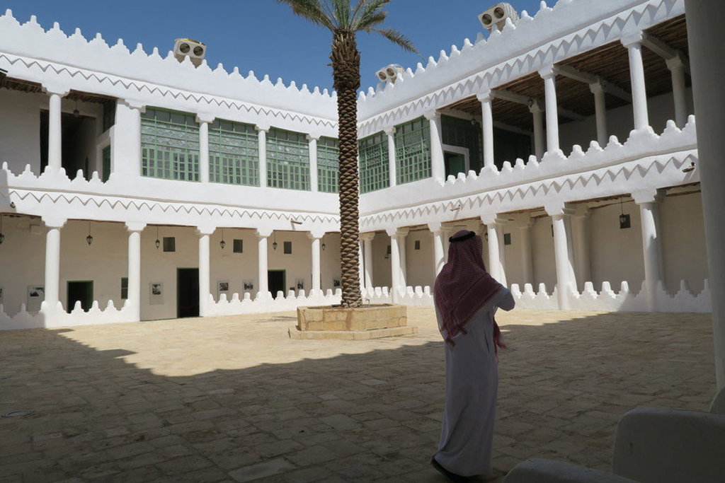 Binnenplaats van historisch fort in Saoedi-Arabië met witte zuilengalerijen, palmboom en bezoeker in traditionele kleding.