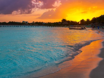 Zonsondergang aan het strand van Isla Holbox, Mexico, met palmbomen en een boot in de branding.