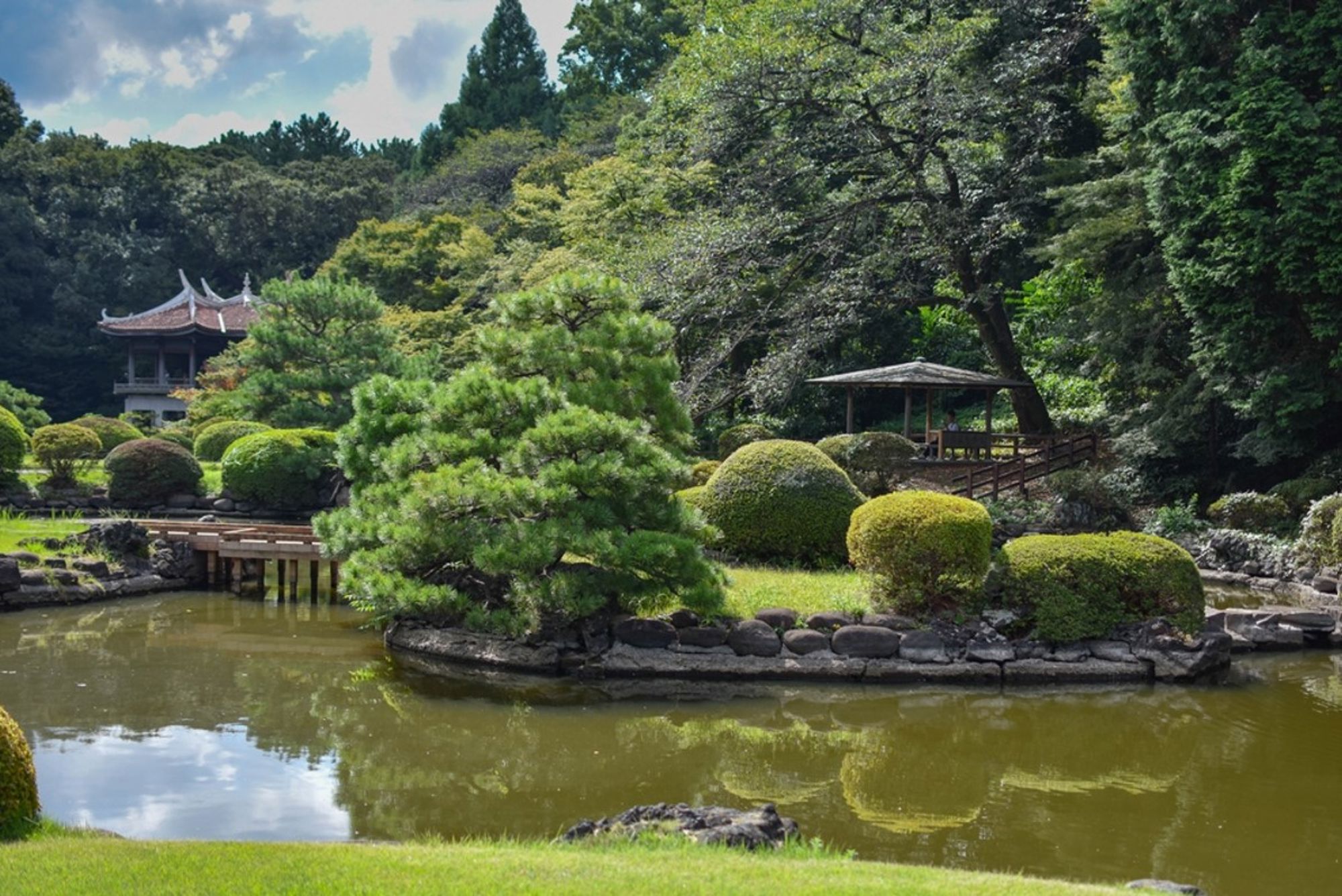 Japanse tuin met vijver, brug en paviljoen tussen groen en gesnoeide struiken.
