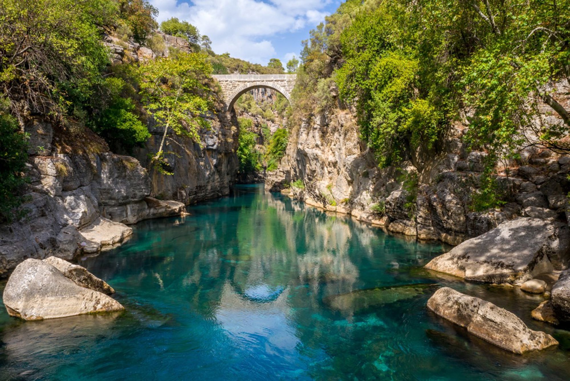 Stenen boogbrug over een smalle kloof met helderblauw water, omringd door rotsen en groen in Turkije.