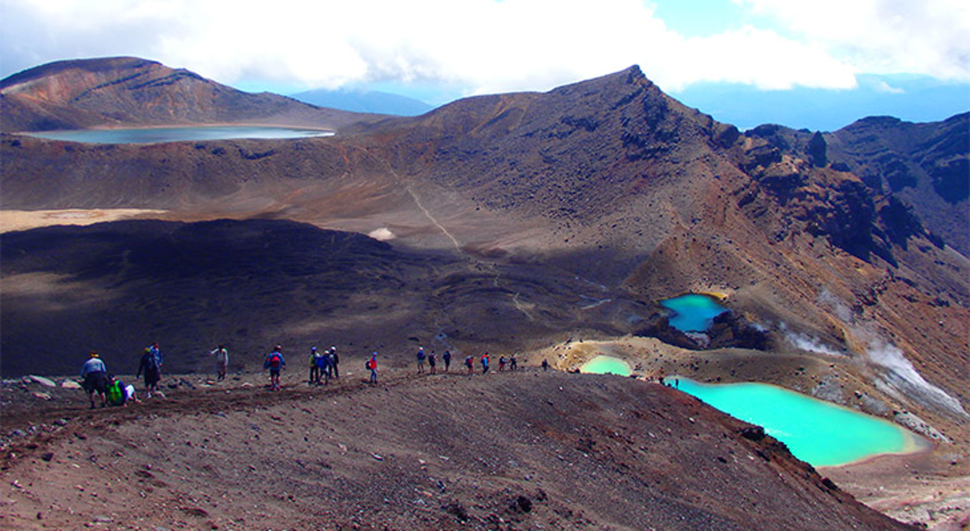 Tongariro Alpine Crossing Nieuw-Zeeland Wandelaars langs een vulkaanlandschap met felblauwe kratermeren en ruige bergen onder een bewolkte lucht.