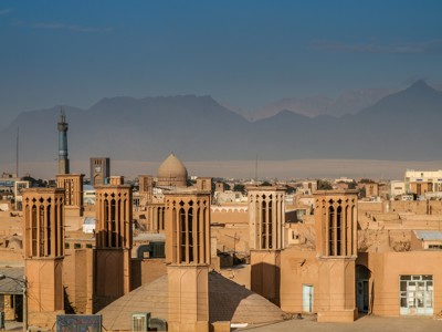 Windtorens en lemen daken in Yazd, met minaret en bergen op de achtergrond.