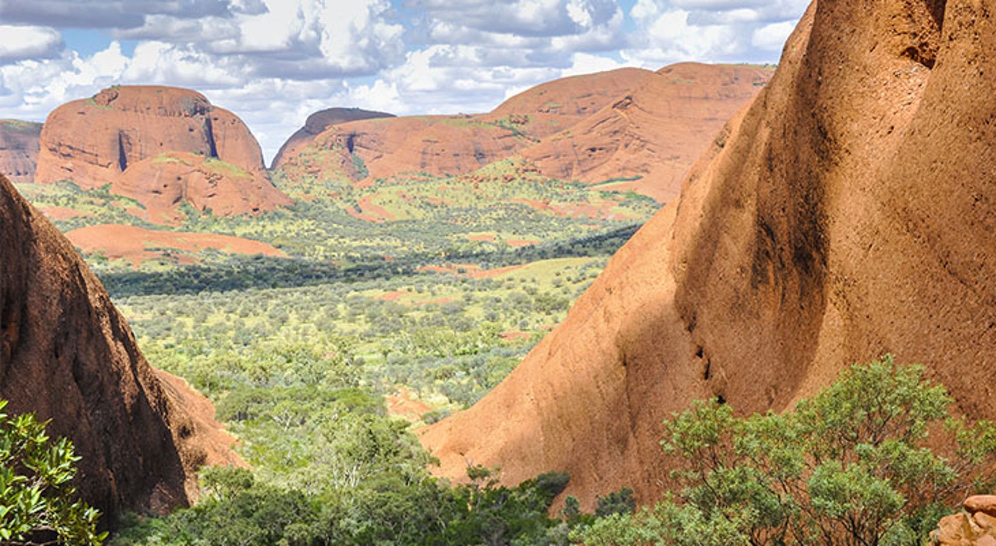 Rode rotsformaties en groene vallei in Kata Tjuta, Australië, onder een blauwe lucht met wolken.