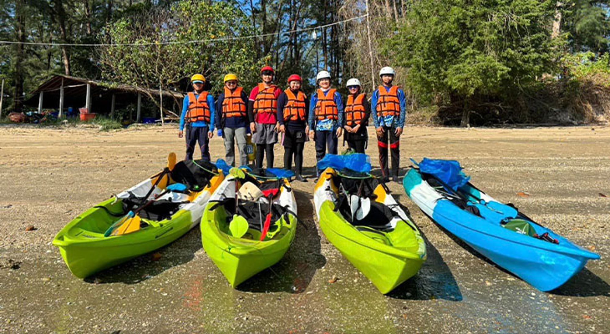 Groep kajakkers met helmen en reddingsvesten op het strand, met kajaks klaar voor vertrek.