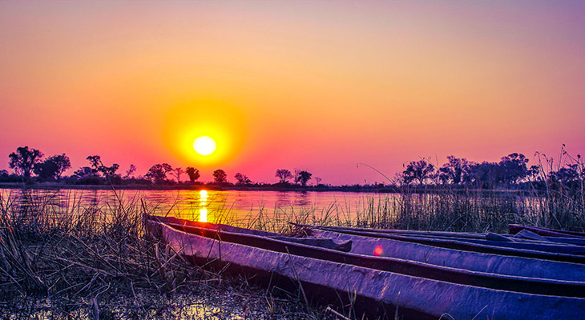 Zonsondergang boven een rustige rivier met kano’s aan de oever en silhouetten van bomen aan de horizon.