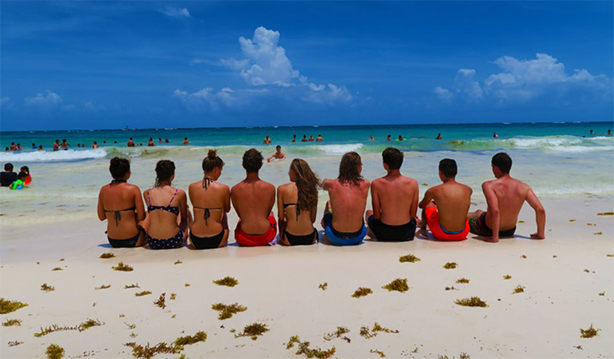 Samen tijd doorbrengen op het strand Groep jongeren zit op het strand en kijkt uit over de zee met zwemmers en wolkenlucht.