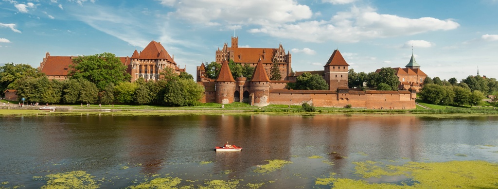 Kasteel Malbork aan de rivier de Nogat in Polen, met rode daken en stadsmuren onder een blauwe lucht.