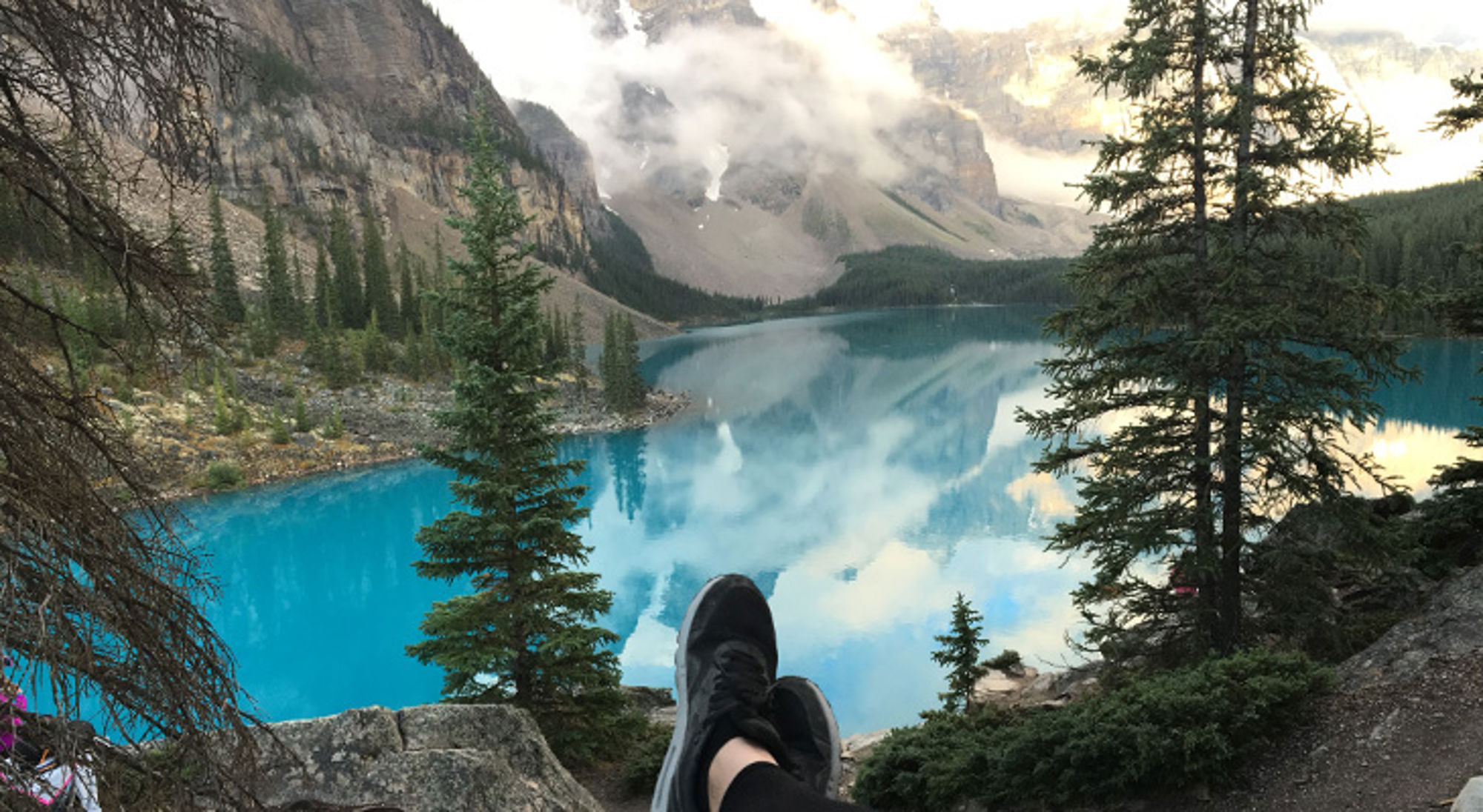 uitzicht op meer in canada op singlereis Uitzicht op een turquoise bergmeer met dennenbomen en mistige bergtoppen, gezien vanaf een rots.