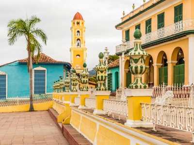 Kleurrijke straat in Trinidad, Cuba, met gele kerktoren, palmbomen en sierlijk hekwerk.