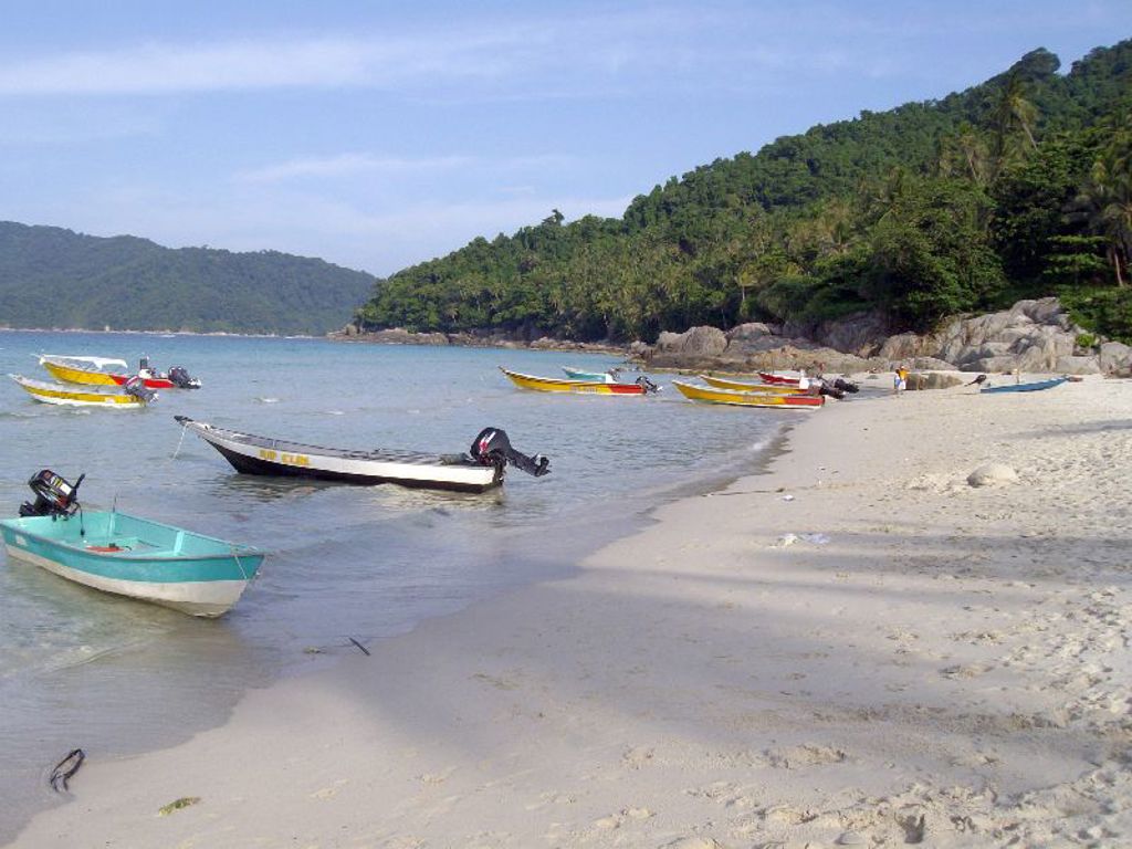 Strand in Maleisië met kleurrijke bootjes voor de kust en groene heuvels op de achtergrond.