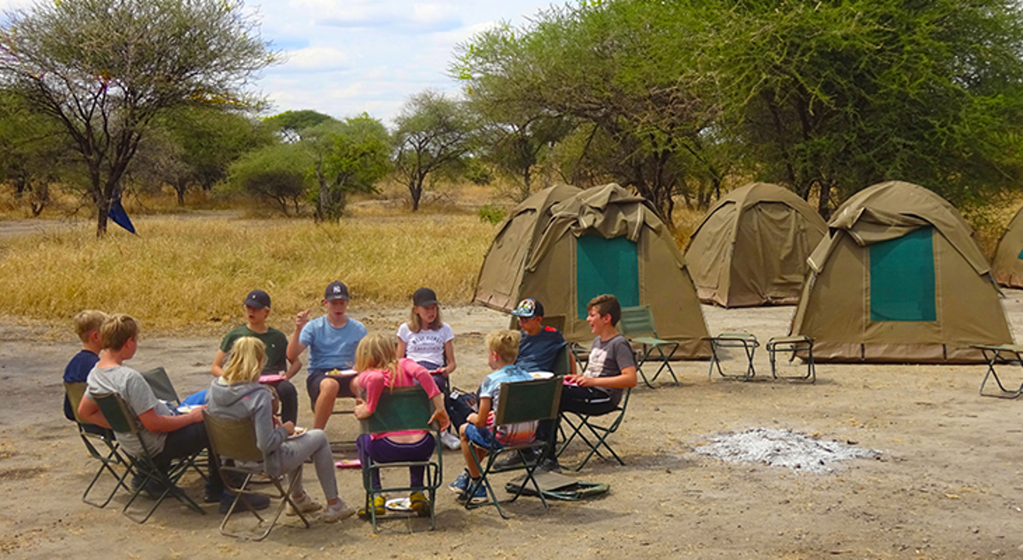Kinderen zitten in een kring op campingstoelen bij safaritenten op een kampplaats in de savanne.