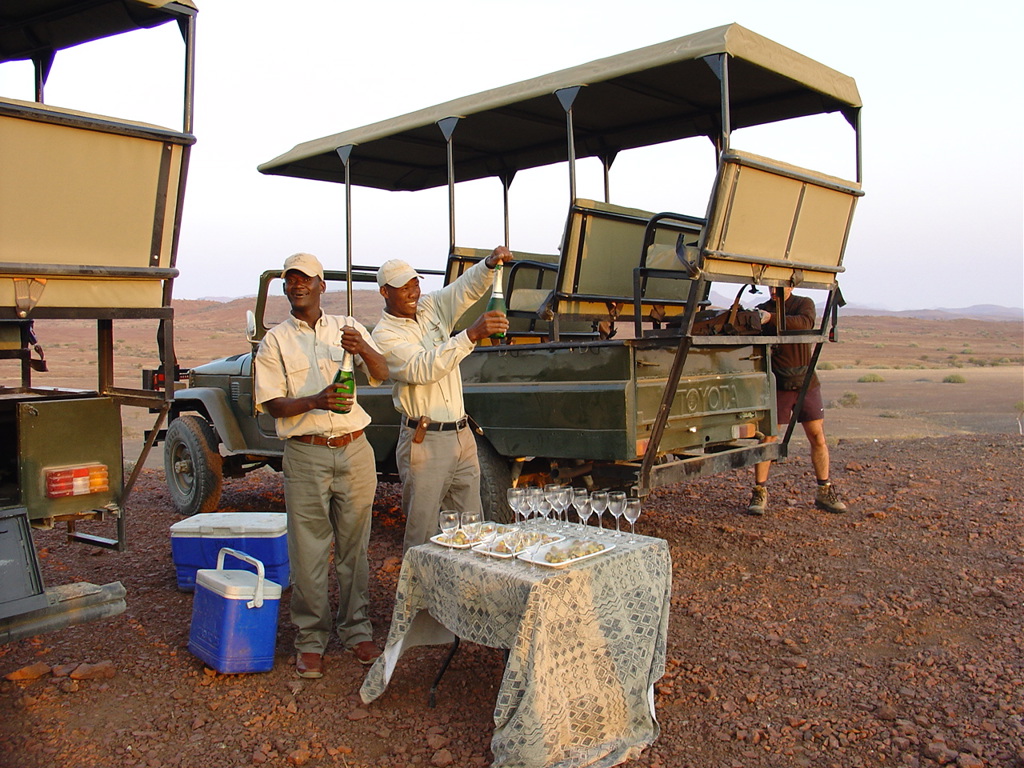 Safari-gidsen schenken champagne bij sundowner naast safaritruck tijdens groepsrondreis Namibië lodge/hotel.