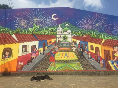 Muurschildering van Ataco met kleurrijke straat, kerk en vuurwerk, op de route Guatemala, El Salvador en Honduras.
