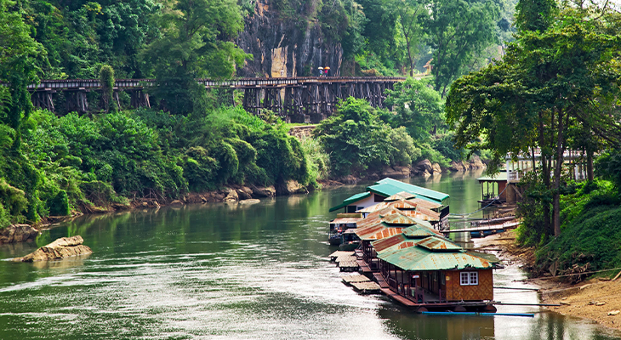 River Kwai Thailand Drijvende huisjes op een rivier, met groene jungle en een houten brug op de achtergrond.