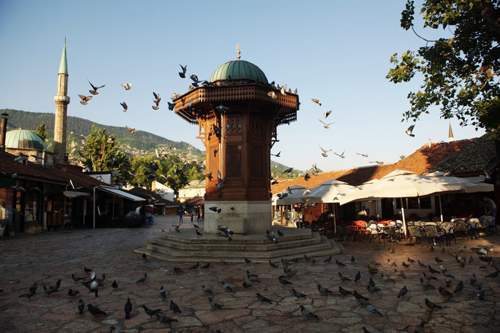 Houten fontein Sebilj op het Baščaršija-plein in Sarajevo, met duiven en minaret op de achtergrond.