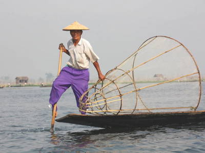 Visser op het Inlemeer in Myanmar, staand op een boot en roeiend met een been naast een groot visnet.