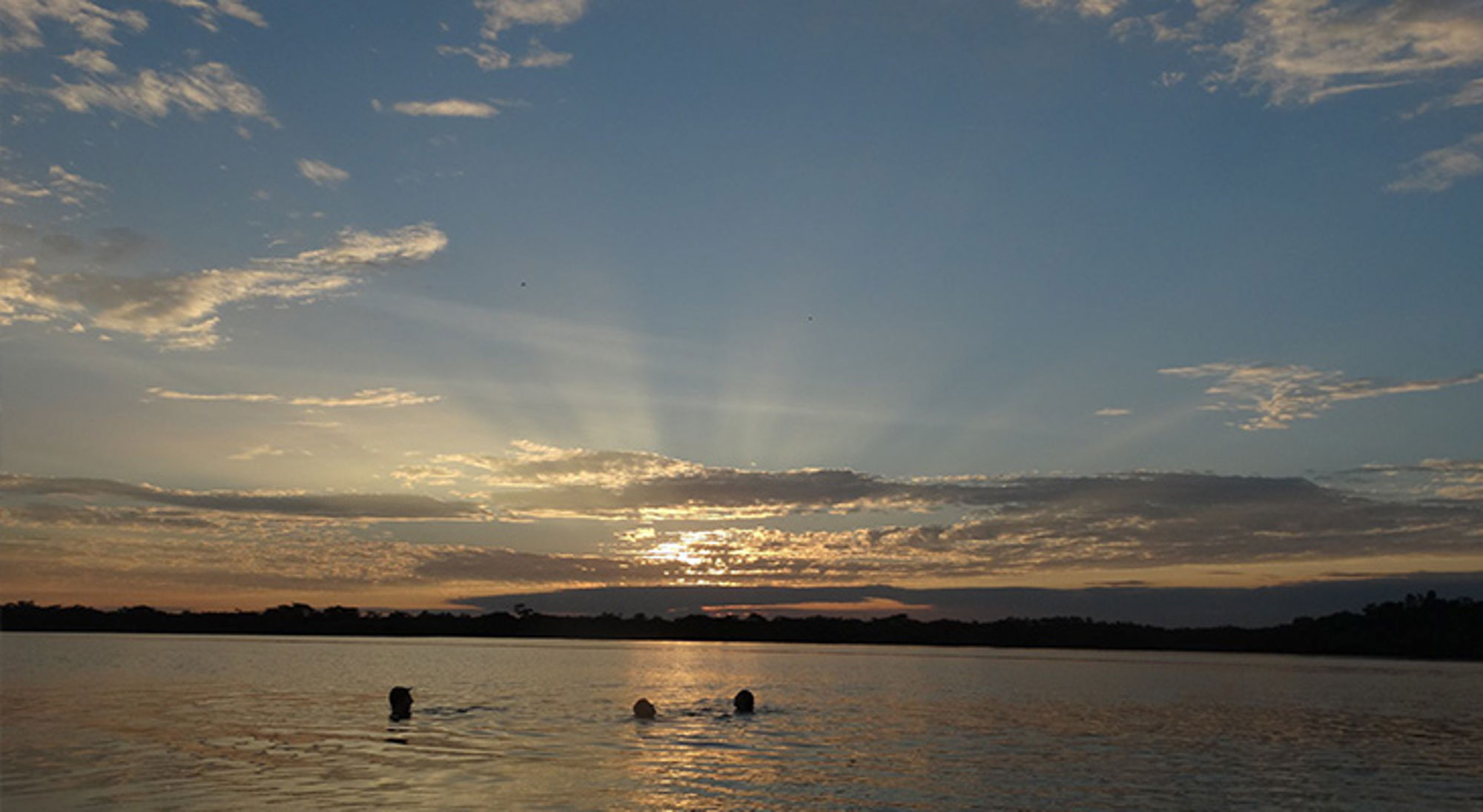 Laguna Grande in Ecuador Zonsondergang boven een rivier, met drie zwemmers in het water en wolkenluchten aan de horizon.