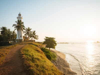 Vuurtoren aan de kust van Sri Lanka met palmbomen en strand bij zonsondergang.