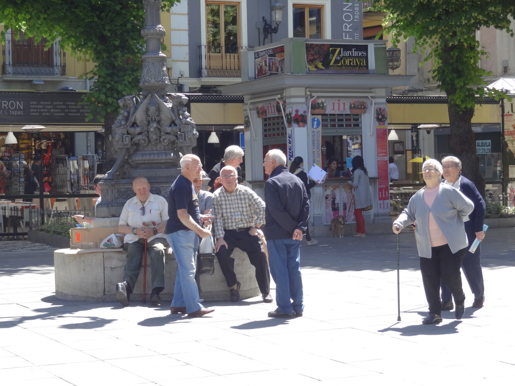 Mensen op een zonnig plein in Andalusië met fontein en terrasjes op de achtergrond.
