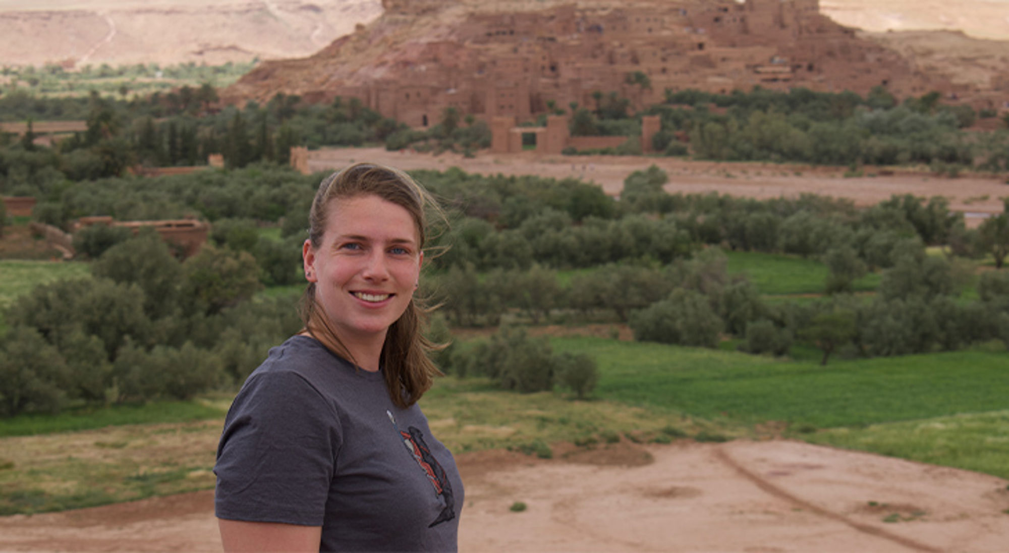 Carolien in Marokko Vrouw poseert met op de achtergrond een groene vallei en een lemen kasbah in een berglandschap.