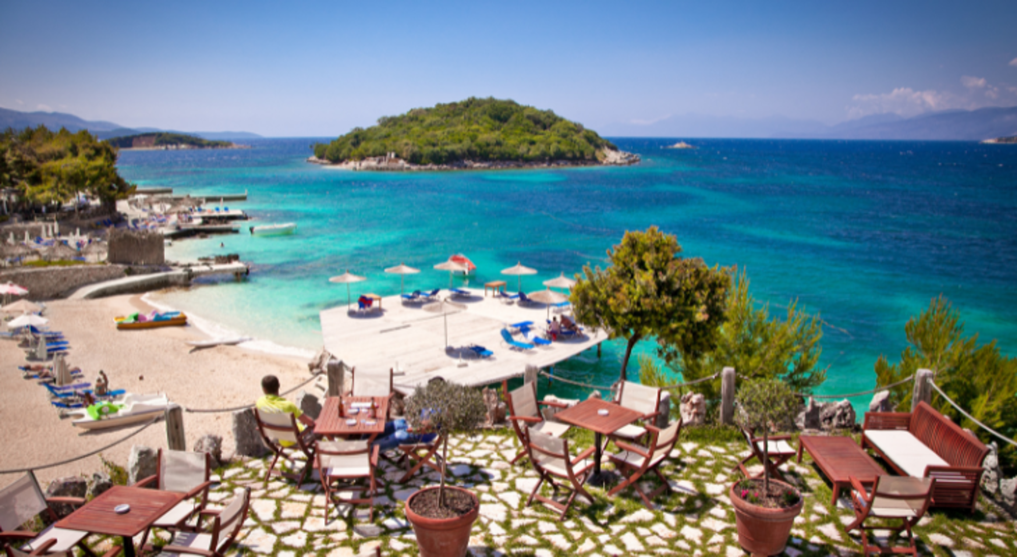 Terras met tafels en stoelen met uitzicht op een turquoise baai, zandstrand en groen eiland.