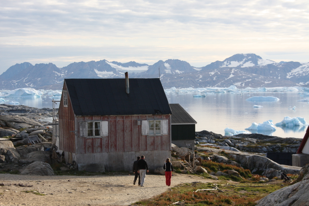 Huis aan de kust bij Disko Bay in Groenland, met ijsbergen in zee en bergen op de achtergrond.