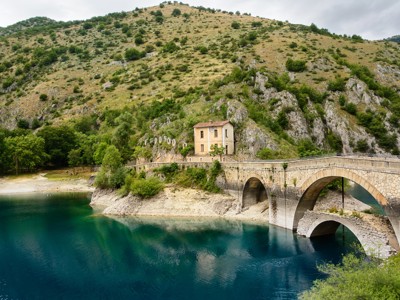 Stenen boogbrug met huisje aan een turquoise bergmeer in de Centrale Apennijnen, Italië.
