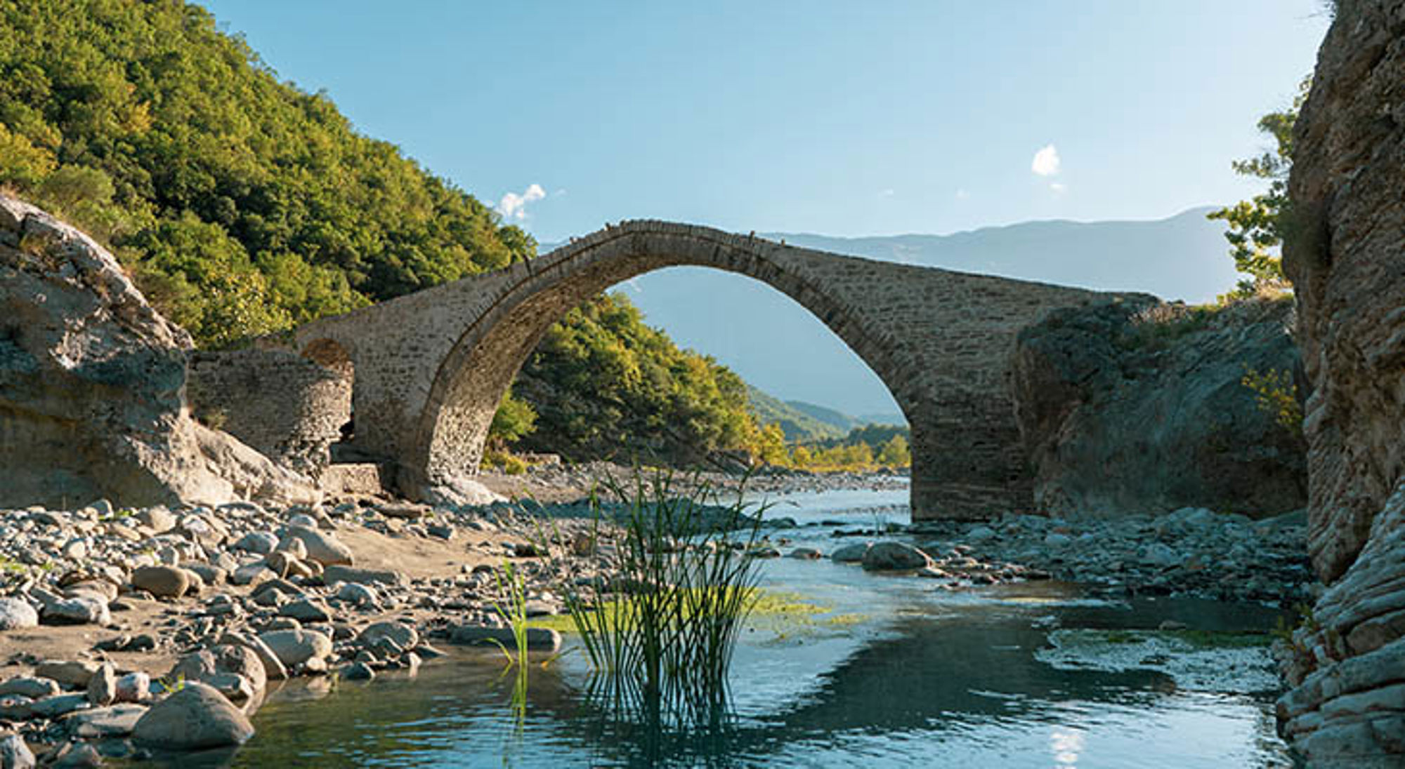 Permet Albanië Oude stenen boogbrug over een ondiepe rivier tussen groene heuvels en rotsen.