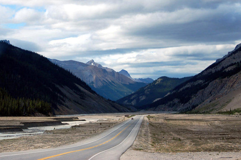 Lange weg door een bergvallei in West-Canada, met rivierbedding en besneeuwde toppen onder wolkenlucht.