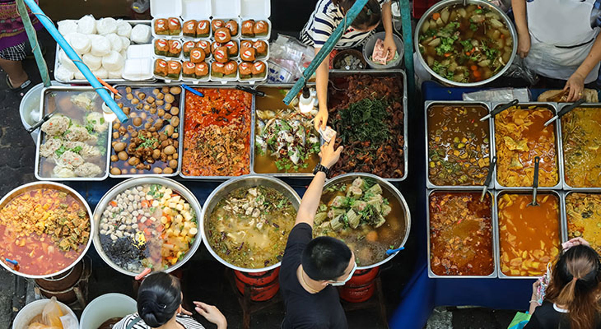 Bovenaanzicht van een streetfoodkraam met grote pannen soep, curry en snacks op een lokale markt.