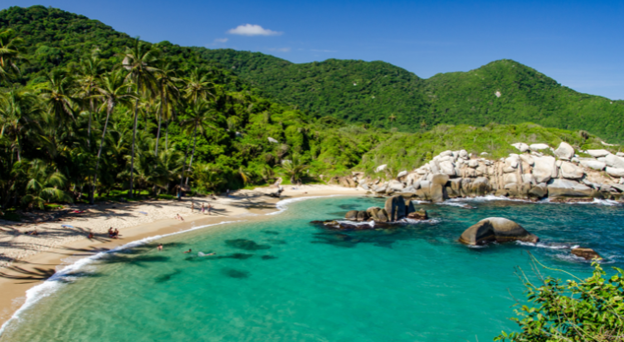 Tropische baai met wit zandstrand, palmbomen, turquoise zee en rotsen, omringd door groene heuvels.