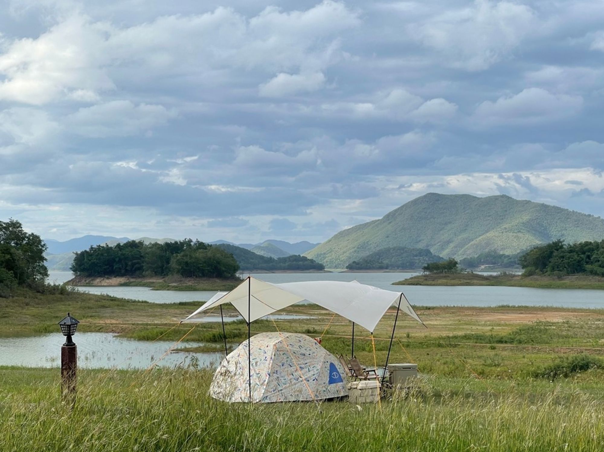 Kampeerplek met tent en luifel aan een meer, met groene bergen op de achtergrond in Thailand.