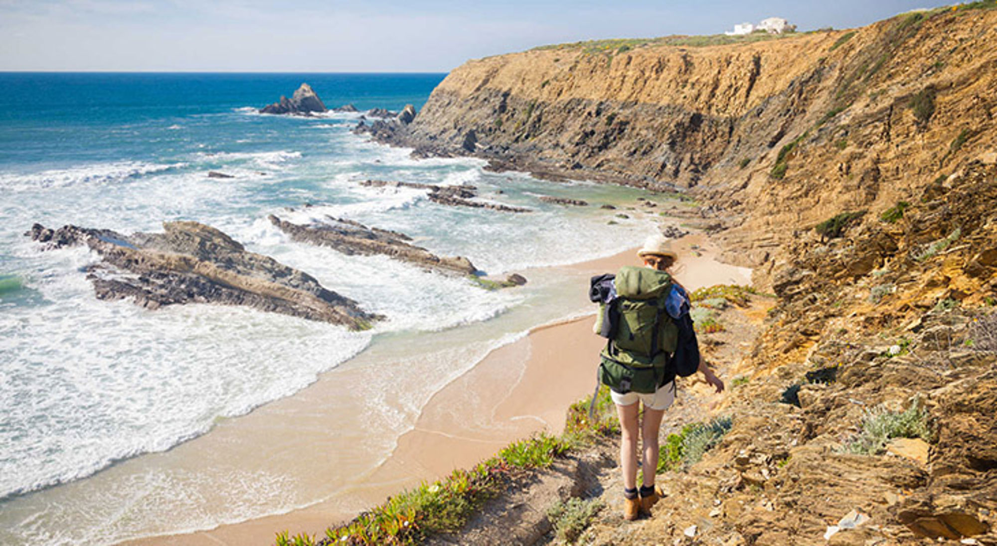 Wandelvakantie Portugal Wandelaar met rugzak op klifpad langs ruige kust met strand en golven.