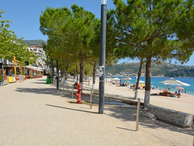 Boulevard langs het strand in Albanië met pijnbomen, terrasjes en parasols aan zee.