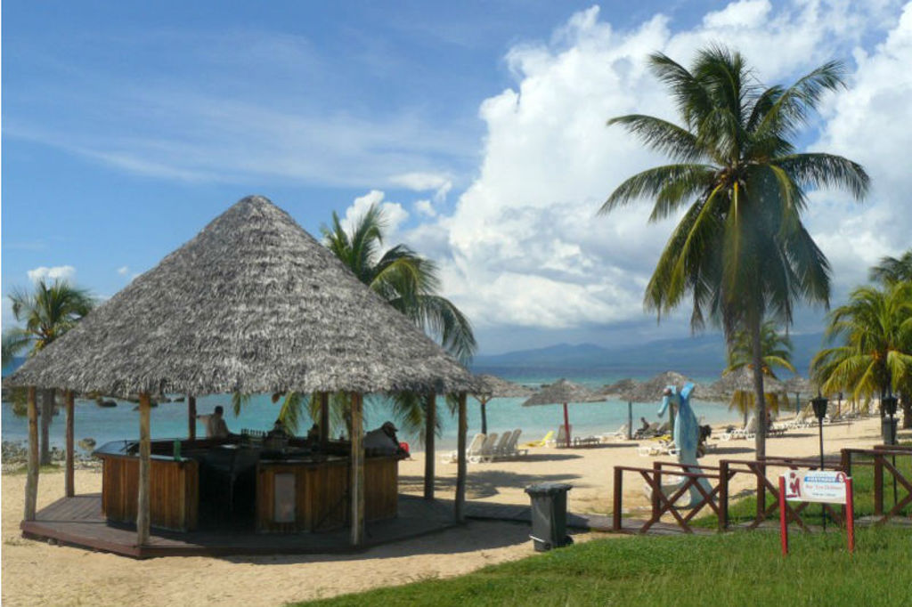 Strand in Cuba met rieten strandbar, palmbomen en ligbedden aan helderblauwe zee.