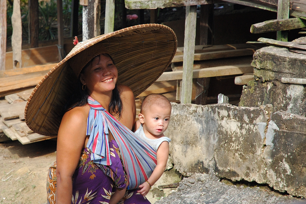 Vrouw met grote hoed draagt baby in draagdoek in een dorp op Maleisisch Borneo.