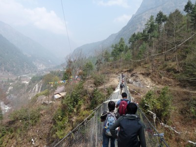 Wandelaars steken een hangbrug over tijdens de Route Nepal Everest Base Camp, met bergvallei en gebedsvlaggen.