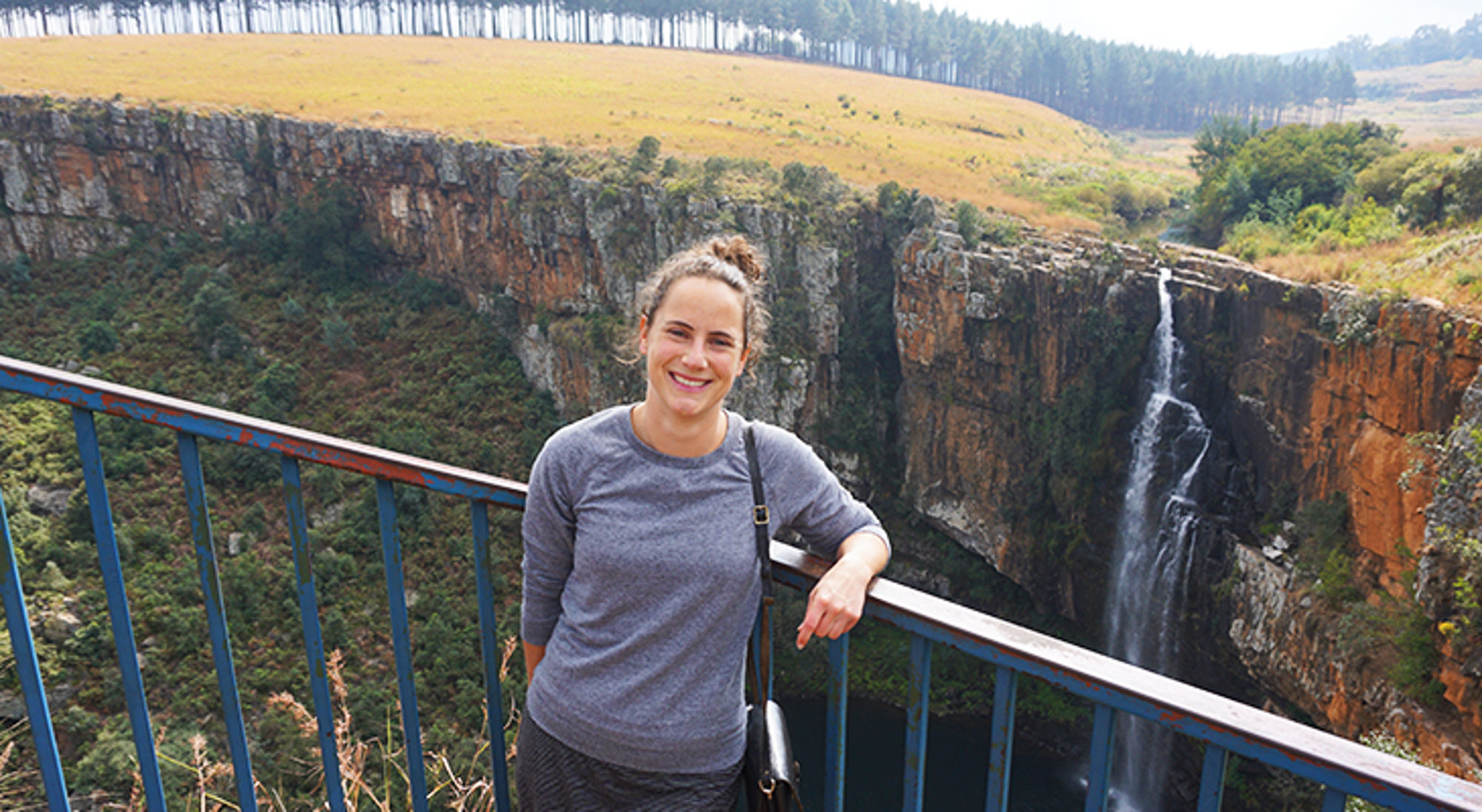 Vrouw poseert bij uitkijkpunt met hoge waterval in een kloof op de achtergrond.