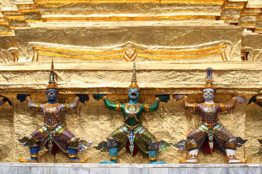Gekleurde tempelwachters in mozaïek tegen een gouden muur bij een boeddhistische tempel in Thailand.