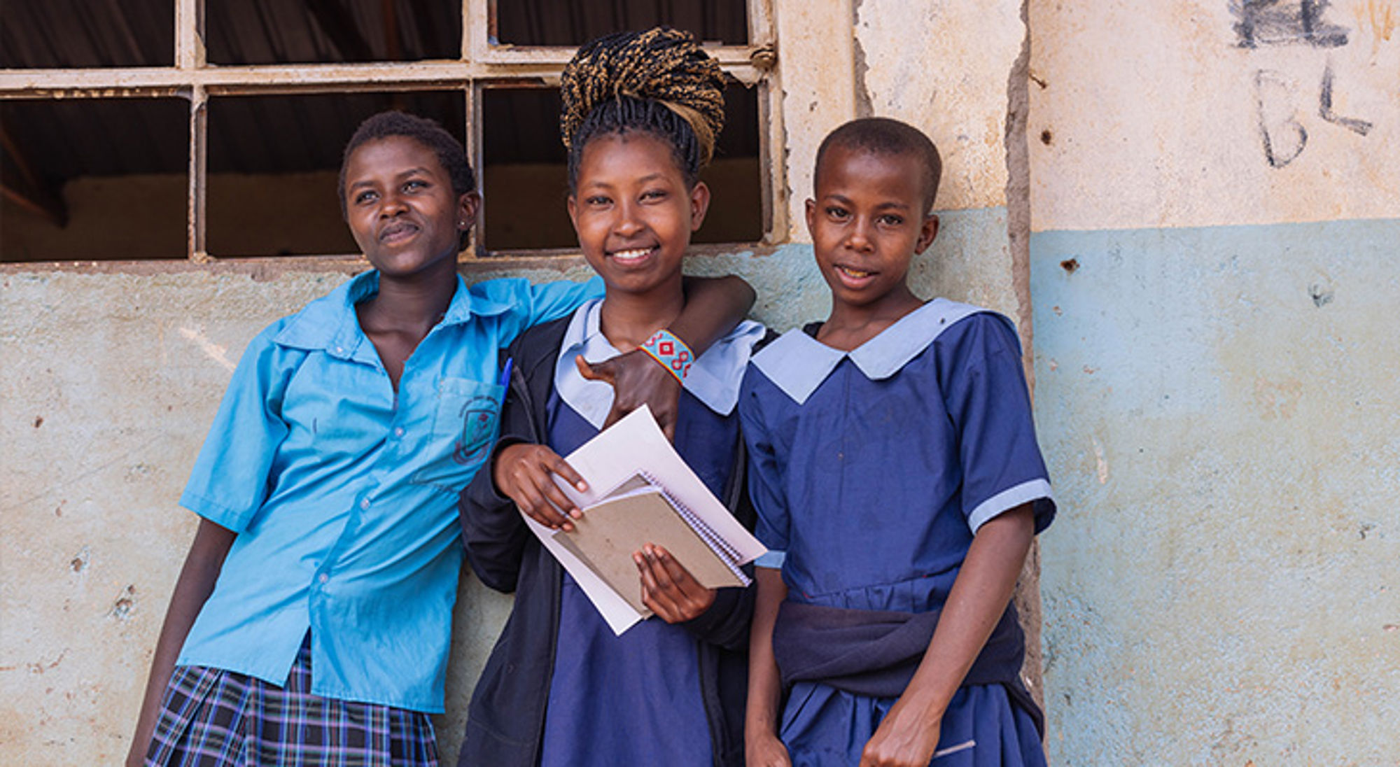 Drie scholieren in schooluniform poseren met schriftjes voor een schoolgebouw.