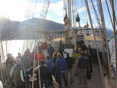 Reizigers op het dek van een zeilschip tijdens een vaartocht, met bergen op de achtergrond.