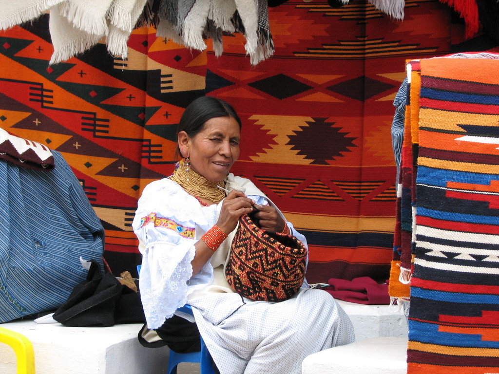 Vrouw in traditionele klederdracht maakt handwerk op een kleurrijke markt in Ecuador.