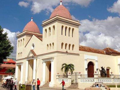 Kathedraal met twee roze koepels in Santiago de Cuba, met mensen op straat en een oude auto op de voorgrond.
