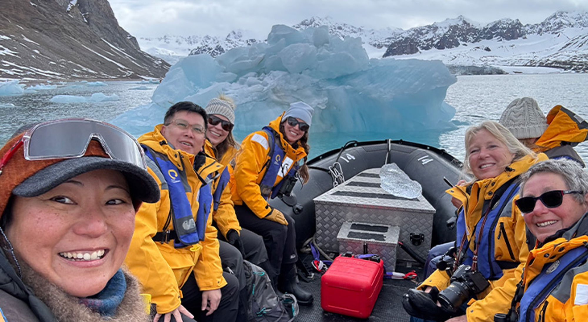 Zodiac Groep reizigers in zodiacboot met gele jassen bij een ijsberg in een fjord met besneeuwde bergen.