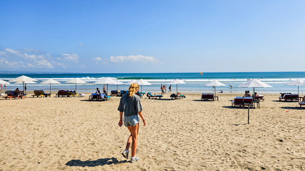 Strand op Bali met ligbedden en parasols, terwijl een reiziger richting de zee loopt.